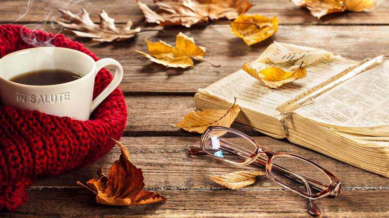 Su un tavolo di legno ci sono delle foglie d'acero gialle, una tazza di tè avvolta da una sciarpa rossa, un libro aperto e un paio di occhiali da vista