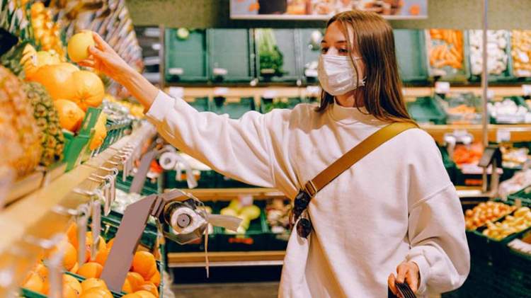 Una ragazza con la mascherina mentre prende un'arancia dallo scaffale del supermercato