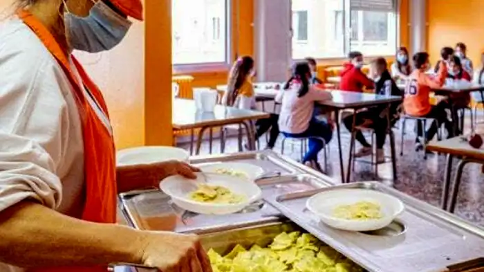 L'alimentazione a scuola durante il Covid. Viene raffigurata una mensa scolastica con in primo piano la cuoca che riempie i piatti nei vassoi e sullo sfondo i bimbi seduti a mangiare