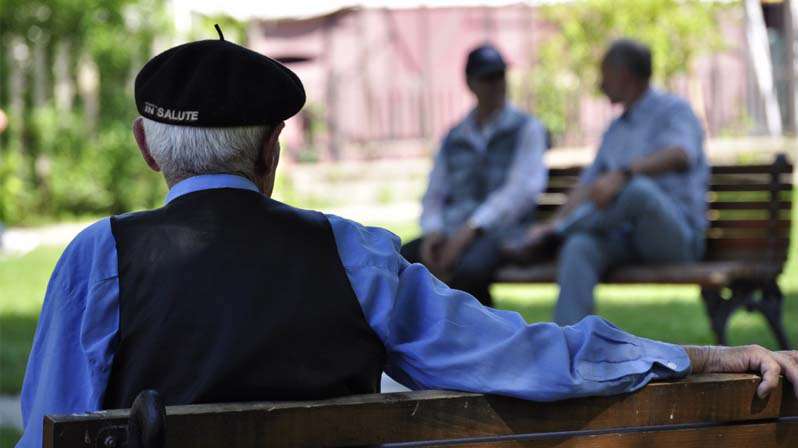 Anziano, di spalle, seduto su una panchina al parco guarda padre e figlio che parlano seduti su una panchina di fronte a lui 