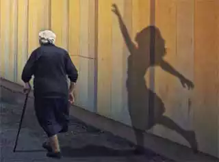 Donna anziana che passeggia con un bastone mentre sul muro, alla sua destra, la sua ombra giovane balla