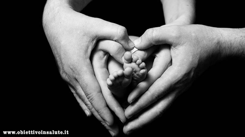 Mani del papà disposte a cuore che contengono quelle della madre sempre disposte a cuore all'interno delle quali ci sono i piedini del figlio