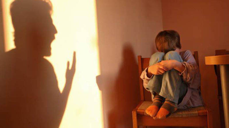 Un bambino rannicchiato su una sedia con la fronte sulle ginocchia, mentre sul muro si vede l'ombra del padre che urla