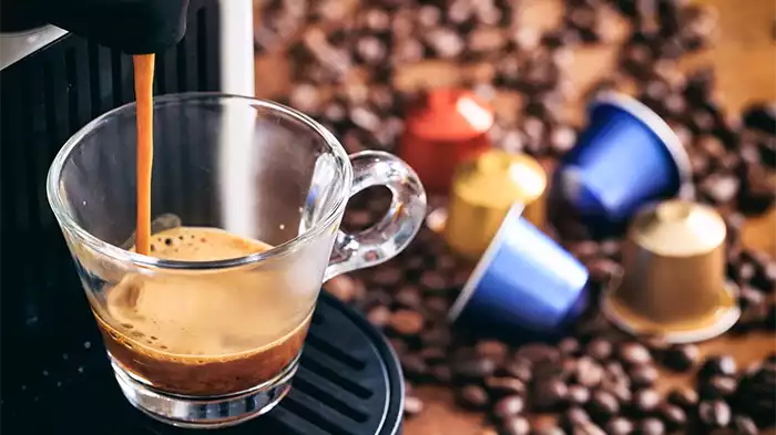 In primo piano una macchinetta per il caffè in funzione e sullo sfondo dei chicchi di caffè con delle capsule sul tavolo