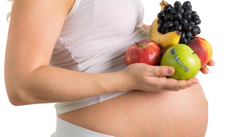 Donna incinta ha, nelle mani, della frutta (mele, pesca, uva e banana)
