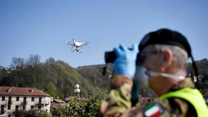 Carabiniere con la mascherina controlla con un binocolo, mentre in cielo vola un drone